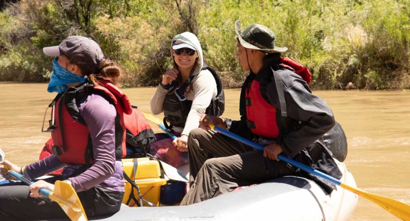 Three people wearing life jackets and hats sit aboard a raft on calm water. 